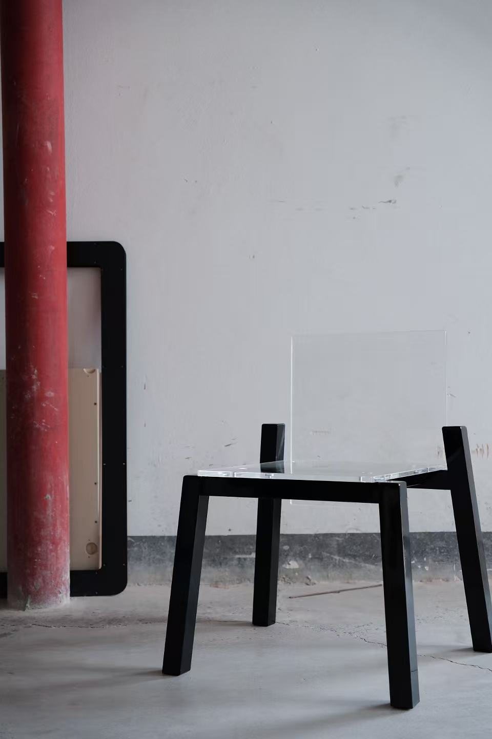 Float Chair with black legs and a clear acrylic seat, against a concrete wall beside a red column